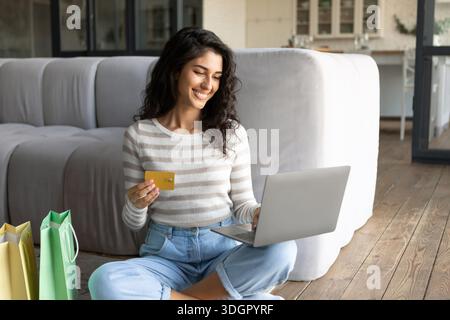 Cybershopping. Giovane signora caucasica con computer portatile, borse shopping e carta di credito che acquista beni nel negozio online a casa Foto Stock