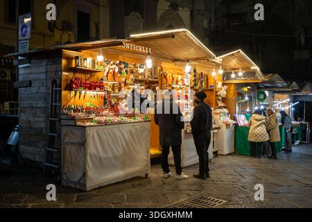 Napoli, Italia - 26 dicembre 2025: Le persone si sono riunite intorno a un mercatino di Natale in legno di notte, vendendo tradizionali ciondoli rossi fatti a mano e locali Foto Stock