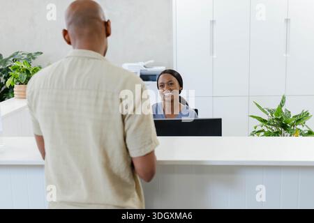 Receptionist afroamericano seduto alla scrivania in cima blu, visitatore maschio in piedi vicino alla stampante Foto Stock