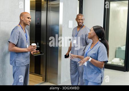 Lavoratori afroamericani in scrub in piedi e chiacchierare nella hall dell'ascensore tenendo il telefono con caffè Foto Stock