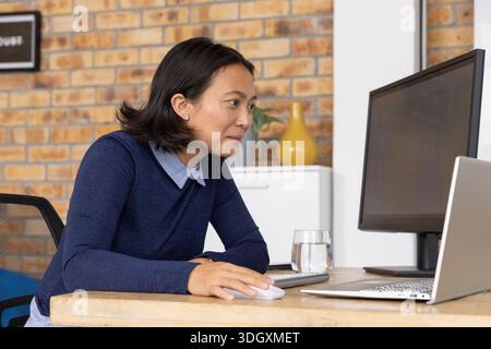 Donna asiatica che si appoggia in avanti alla scrivania dell'ufficio e indossa un maglione navy usando il mouse vicino al monitor e al vetro Foto Stock
