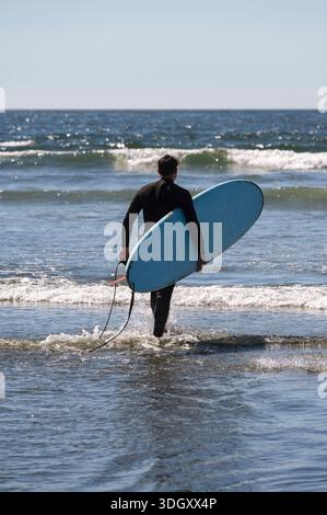 Un uomo in muta che cammina in acqua pronto a fare surf sulle onde dell'oceano a Tofino Canada Foto Stock