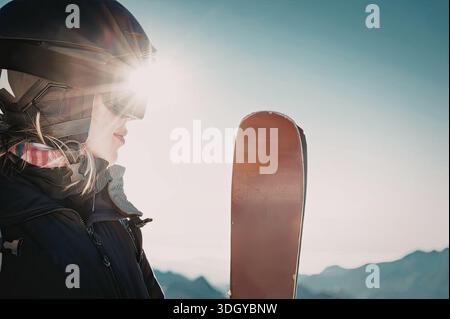 Uno snowboarder, vestito con l'equipaggiamento invernale, si erge con il suo snowboard sullo sfondo di montagne sotto un cielo luminoso. Foto Stock