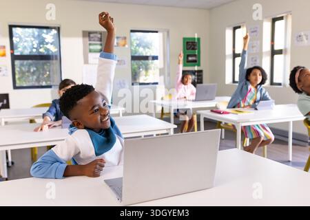 Bambini diversi seduti in classe usando un computer portatile e alzando le mani alle scrivanie bianche con poster Foto Stock