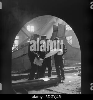 Slaviansk, RSS Ucraina, Unione Sovietica, 1975. Una straordinaria fotografia d'archivio scattata all'interno dello stabilimento di macchinari pesanti di Slavtyazhmash, con un gruppo di quattro ingegneri e lavoratori che discutono di un progetto tecnico. Vista attraverso un arco scuro in primo piano, la scena è illuminata da spettacolari pozzi di luce solare che fluiscono nell'officina. Questa composizione evidenzia la collaborazione tra ingegneria e lavoro manuale durante l'assemblaggio di enormi attrezzature industriali, catturando lo spirito di produzione nella pacifica regione del Donbas. Foto Stock