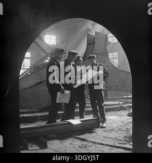 Slaviansk, RSS Ucraina, Unione Sovietica, 1975. Una straordinaria fotografia d'archivio scattata all'interno dello stabilimento di macchinari pesanti di Slavtyazhmash, con un gruppo di quattro ingegneri e lavoratori che discutono di un progetto tecnico. Vista attraverso un arco scuro in primo piano, la scena è illuminata da spettacolari pozzi di luce solare che fluiscono nell'officina. Questa composizione evidenzia la collaborazione tra ingegneria e lavoro manuale durante l'assemblaggio di enormi attrezzature industriali, catturando lo spirito di produzione nella pacifica regione del Donbas. Foto Stock