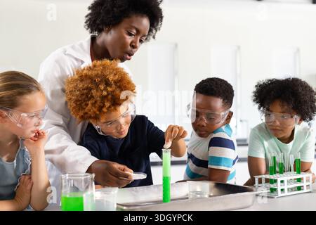 Donna afroamericana che guida i bambini che maneggiano provette verdi al banco del laboratorio indossando camice da laboratorio e occhiali di protezione Foto Stock