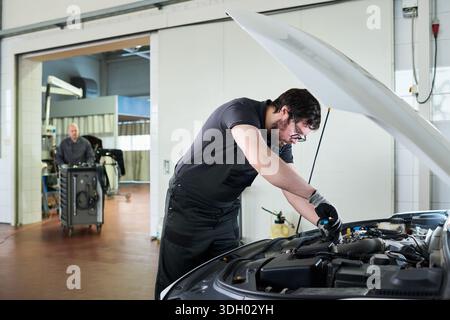 Giovane uomo adulto caucasico che ripara il motore di un'auto in officina, indossa guanti da lavoro e tuta, si concentra su attività meccaniche mentre un uomo caucasico di mezza età sta in piedi in sottofondo osservando Foto Stock