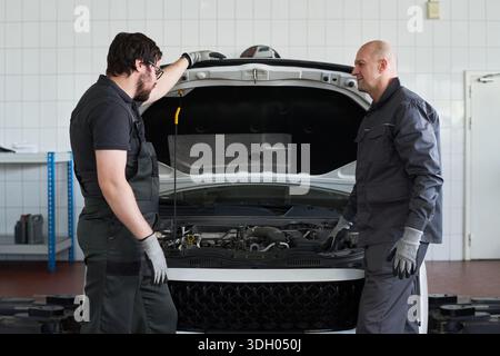Due uomini caucasici di mezza età in piedi di fronte al cofano di un'auto aperta discutono della manutenzione del motore in un'officina di riparazione auto, indossando guanti da lavoro e uniformi meccaniche, si sono concentrati sulla conversazione Foto Stock