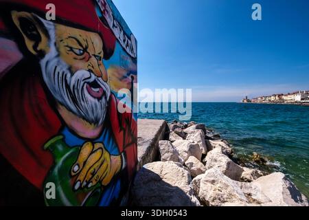 Graffiti colorati su un muro sul mare con la costa rocciosa, gli edifici storici della città, la chiesa di San Clemente e il suo faro in lontananza. Foto Stock