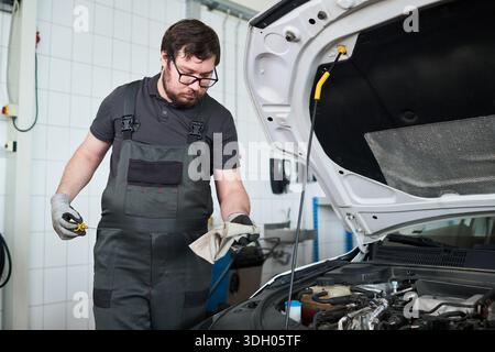 Giovane adulto caucasico che indossa gli occhiali che controlla il livello dell'olio motore sotto il cofano aperto dell'auto in un'officina di riparazione auto, tiene l'astina di livello e la pulisce con un panno, concentrandosi sull'attività di manutenzione Foto Stock