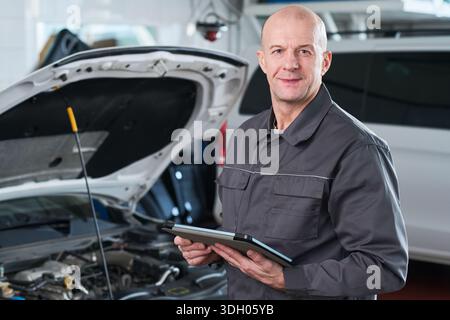 Ritratto di un uomo caucasico di mezza età in piedi in un negozio di riparazione auto con un tablet digitale, sorridendo alla fotocamera con cofano auto aperto in background, dimostrando la diagnostica automobilistica Foto Stock