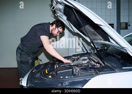 Giovane uomo adulto caucasico che ispeziona il vano motore dell'auto in officina, si appoggia sul cofano aperto ed esamina i componenti meccanici con un'espressione mirata Foto Stock