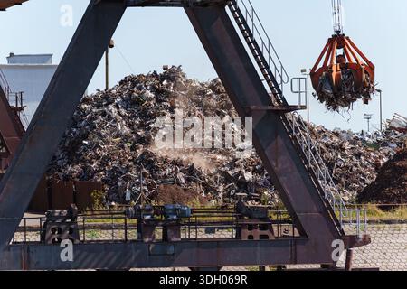 Una massiccia gru solleva i rottami metallici sopra grandi pali vicino agli edifici industriali nell'area del porto Foto Stock