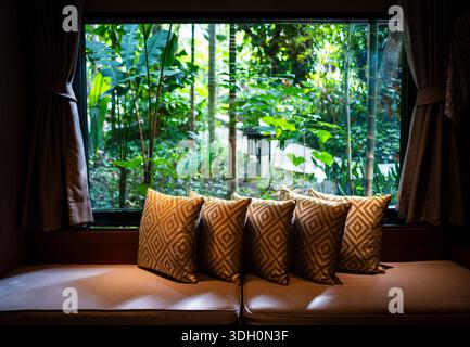 Angolo finestra decorato con cuscini quadrati sul divano con sfondo sfocato del giardino tropicale. Foto Stock