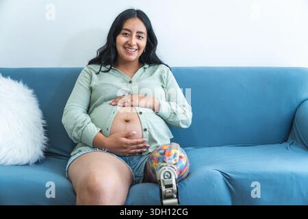 Una donna incinta felice con una gamba protesica è seduta su un comodo divano, abbracciando la sua pancia e sorridendo Foto Stock