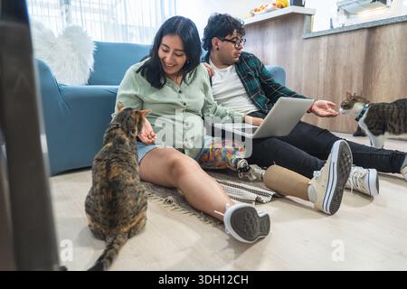 Donna incinta felice con gatto protesico per allattamento alle gambe mentre il suo partner lavora da casa con un laptop Foto Stock