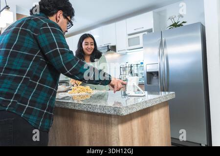 Una giovane coppia prepara l'impasto seguendo la ricetta su un tablet, in una cucina moderna Foto Stock