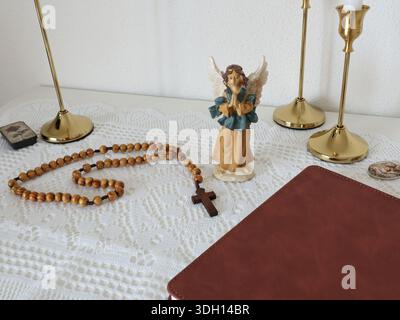Angolo cattolico a casa. Figurina d'angelo, perline di rosario in legno, croce e libro di preghiere. Fede, simboli e tradizioni. Foto Stock