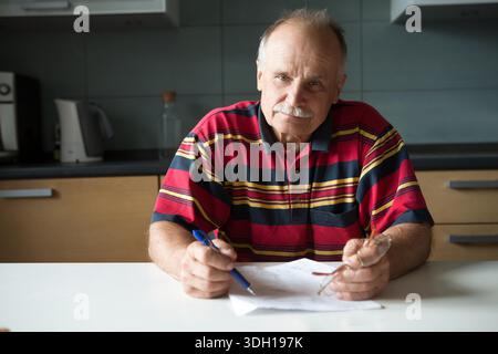 il ritratto dell'anziano seduto a un tavolo in cucina. Un uomo anziano che lavora, paga a casa in cucina. || modello rilasciato Foto Stock