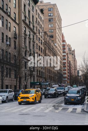 Un taxi giallo di New York guida lungo una strada di Midtown Manhattan costeggiata da auto parcheggiate, alberi senza foglie e condomini. Foto Stock