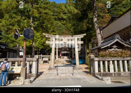 Kinosaki, Giappone - 9 novembre 2023: Una porta torii in pietra si trova all'ingresso di un santuario shintoista, con una tradizionale struttura in legno, recinzione in pietra, alberi e una persona che legge un tabellone informativo visibile sotto la luce del sole. Foto Stock