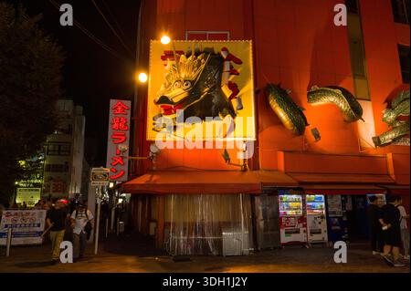 Osaka, Giappone - 5 novembre 2023: Esterno del ristorante Kinryu Ramen a Dotonbori di notte, caratterizzato da un grande cartello con la testa di drago illuminato, elementi scultorei di drago sul muro, segnaletica rossa con scritta giapponese e diversi distributori automatici lungo il marciapiede con persone nelle vicinanze. Foto Stock