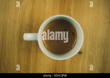 Una tazza di cappuccino vista dall'alto, posizionata su un tavolo di legno, che mostra la schiuma di caffè e una semplice impostazione del piano portapaziente in luce naturale. Foto Stock