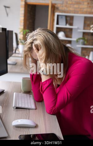 Donna di mezza età che indossa una camicetta rosa seduta alla scrivania che copre il viso con le mani vicino alla tastiera Foto Stock