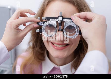 L'optometrista regola il casco di prova durante l'esame di rifrazione per sorridere il paziente in clinica. Adatto per banner di siti Web per la cura degli occhi, annunci pubblicitari e controllo della visione c Foto Stock