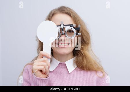 Il paziente sorridente nel telaio di prova copre un occhio con occlusivo durante l'esame di rifrazione. Ideale per siti web di cliniche oculistiche, brochure e annunci per la cura della vista. Foto Stock