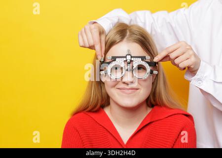 L'optometrista regola il casco di prova per la rifrazione e l'adattamento alla prescrizione durante l'esame oculare con il paziente sorridente su sfondo giallo chiaro Foto Stock
