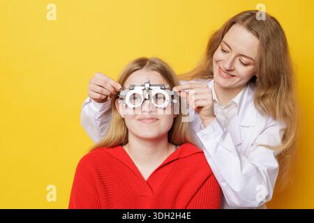 L'oftalmologo sorridente regola la montatura di prova per la rifrazione e la prescrizione di lenti a contatto durante l'esame oculare su sfondo giallo Foto Stock