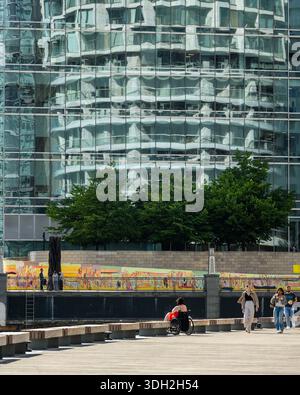 La gente cammina lungo il lungomare di Harbour Quay a Wood Whaf, parte dei Docklands di Londra, con One Park Drive riflesso nelle finestre di 10 Upper Ban Foto Stock