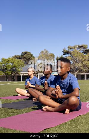 Diversi studenti che indossano magliette blu, si siedono su tappetini da yoga nel campo scolastico, premono le mani Foto Stock