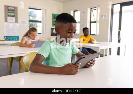 Bambini diversi seduti in classe, tenendo i tablet e lavorando alle scrivanie accanto alle finestre Foto Stock