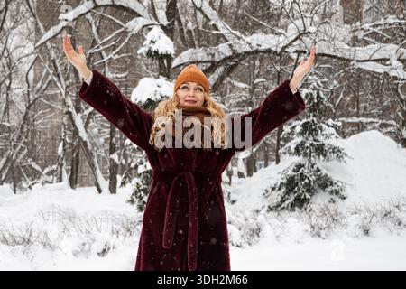 Donna adulta sorridente che alza le braccia e si diverte a scivolare la neve durante la passeggiata invernale nel parco nevoso della città. Donna che indossa un caldo cappotto, sciarpa e cappello Foto Stock