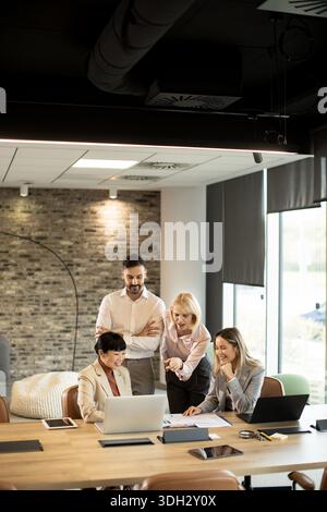 Quattro colleghi si riuniscono intorno a un notebook in un elegante ufficio. Discutono le idee e condividono informazioni utili per migliorare i risultati dei progetti in un Foto Stock