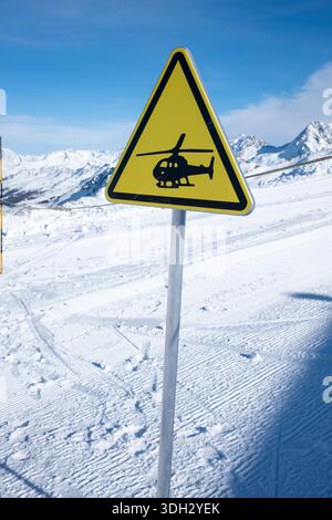 Segnale di avvertimento giallo e nero di un'area di atterraggio per elicotteri, cima montuosa innevata contro il cielo blu chiaro, un paesaggio invernale della Francia Foto Stock