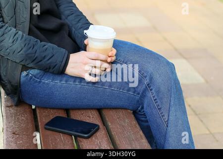 Visione mirata del latte da asporto a mano sulla panca in legno accanto allo smartphone spento in ambienti esterni. Foto Stock