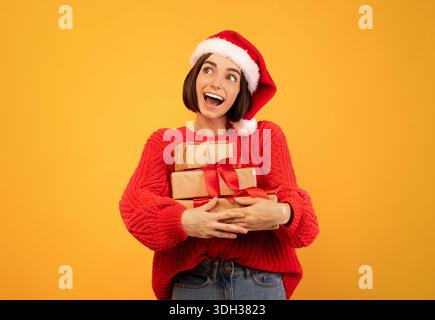 Concetto di vendita per le vacanze. Signora gioiosa in santa cappello tenendo stack di regali di Natale, guardando da parte su sfondo giallo Foto Stock