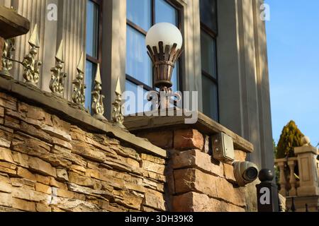 Tradizionale luce a lampione in ferro battuto con luce bianca soffusa su base in pietra accanto alla classica facciata a colonne, con telecamera a cupola per la sicurezza. Foto Stock
