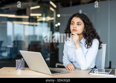 Giovane donna d'affari con capelli scuri ricci seduta a una scrivania con un notebook, profondamente coinvolta nel pensiero e concentrata sulle sue ambizioni professionali e sul suo successo Foto Stock
