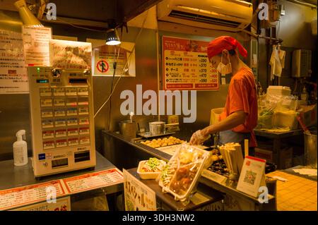 Kyoto, Giappone - 18 ottobre 2023: Un membro del personale che indossa un berretto rosso e una maschera facciale prepara takoyaki su una griglia in una bancarella alimentare nel mercato di Nishiki, con un distributore automatico, cartelli scritti a mano e tabelloni da menu visibili. Foto Stock