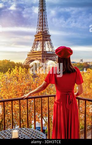 Una bella donna con un abito rosso si erge su un balcone e gode della vista dell'alba autunnale della Torre Eiffel a Parigi, Francia Foto Stock