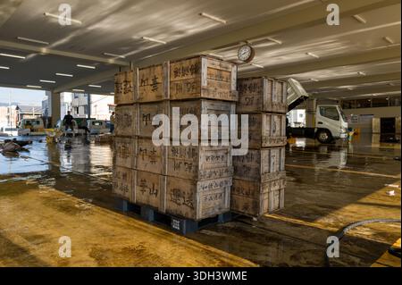 Nachikatsuura, Giappone - 27 ottobre 2023: Pile di casse di legno con scritta giapponese sono disposte su pallet all'interno di un mercato del pesce coperto, con un orologio sul soffitto, pavimenti bagnati e lavoratori e veicoli visibili sullo sfondo. Foto Stock