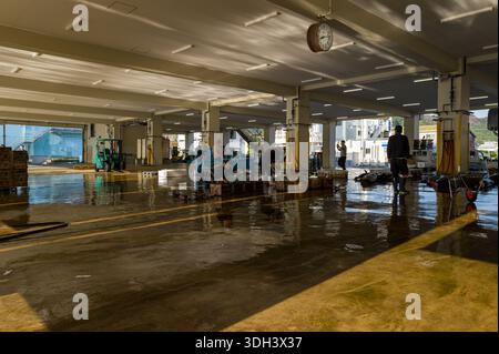 Nachikatsuura, Giappone - 27 ottobre 2023: I lavoratori gestiscono casse e attrezzature su un pavimento di cemento bagnato all'interno di un mercato ittico coperto, con carrelli elevatori, grandi contenitori blu, un orologio a parete e veicoli visibili sullo sfondo. Foto Stock