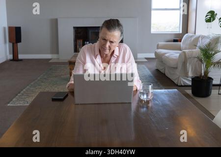 Donna matura in camicia rosa che lavora a casa con un laptop argentato, uno smartphone a sinistra, l'acqua a destra Foto Stock