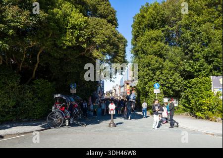 ISE, Giappone - 29 ottobre 2023: Gruppi di persone entrano nel quartiere Okage Yokocho, con i risciò allineati sul lato sinistro, grandi alberi in testa e cartelli visibili sotto il cielo azzurro. Foto Stock