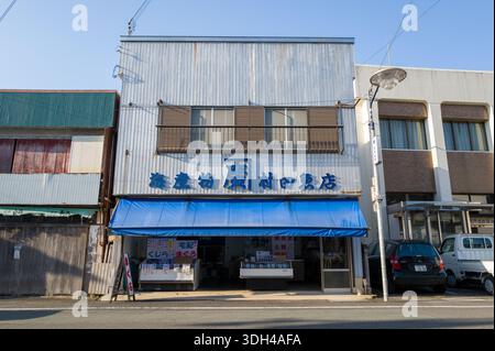 Nachikatsuura, Giappone - 27 ottobre 2023: L'esterno di un negozio di pesce con cartellonistica giapponese e una tenda blu si vede su una strada, fiancheggiata da edifici adiacenti e veicoli parcheggiati. Foto Stock